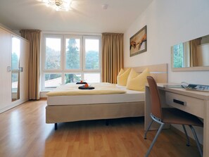Une chambre avec un lit, un bureau et une chaise. Il y a des fenêtres avec des rideaux, un tableau au mur et un luminaire au plafond.