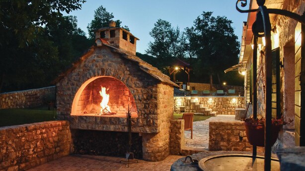 An outdoor stone fireplace with a fire burning inside, surrounded by stone walls and a paved area. - Beautiful home in Hrvace (Hrvace)