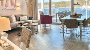 Un salon moderne comprenant un canapé, un fauteuil rouge, une table à manger et des chaises, avec vue sur un front de mer où l’on aperçoit des bateaux et des bâtiments.