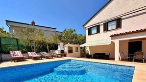 Uma piscina com espreguiçadeiras, área de churrasco e um edifício com telhado de azulejos.