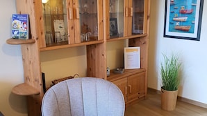 Ein Holzkabinent mit Glastüren, ein grau gepolsterter Sessel, ein gerahmtes Bild an der Wand und ein Blumentopf.