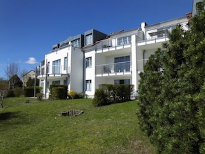 A multi-story residential building with balconies, a well-maintained lawn, and a pine tree.