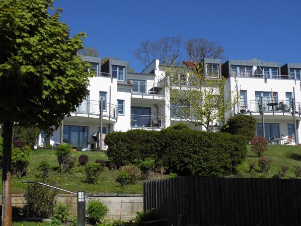 A multi-story building with balconies, surrounded by greenery and a fenced area.
