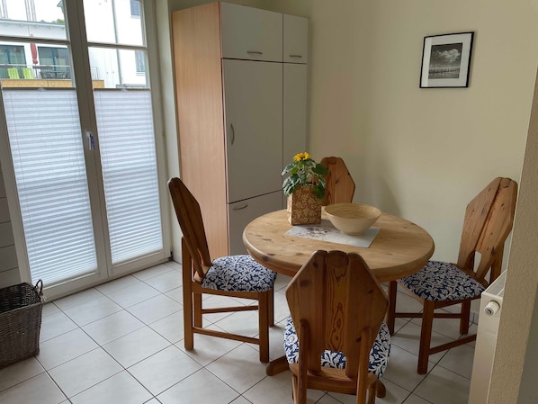 Un coin repas comprenant une table ronde, quatre chaises en bois, un buffet blanc et une fenêtre avec des stores.