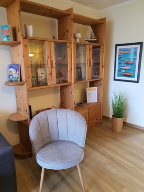 Un meuble en bois avec des portes en verre, un fauteuil recouvert d’un tissu gris, un tableau encadré accroché au mur et une plante en pot.