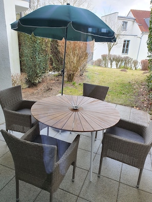 Une table ronde entourée de chaises en osier et surmontée d’un parasol vert foncé.