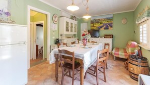 Un coin repas avec une table dressée pour quatre personnes, entourée de chaises en bois. On aperçoit un réfrigérateur blanc sur la gauche, un meuble avec des portes en verre et un tableau accroché au mur.