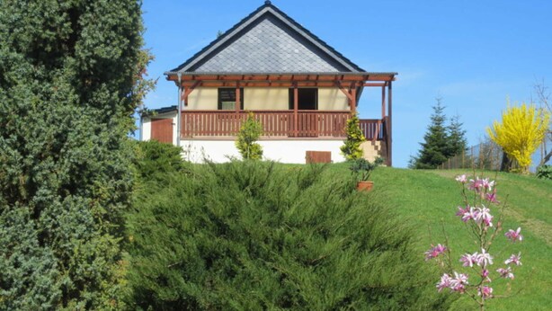 A house with a sloped roof, wooden balcony, and a small garden with flowering plants. - Apartment 1 - Apartment 1 (72389) (Lychen)