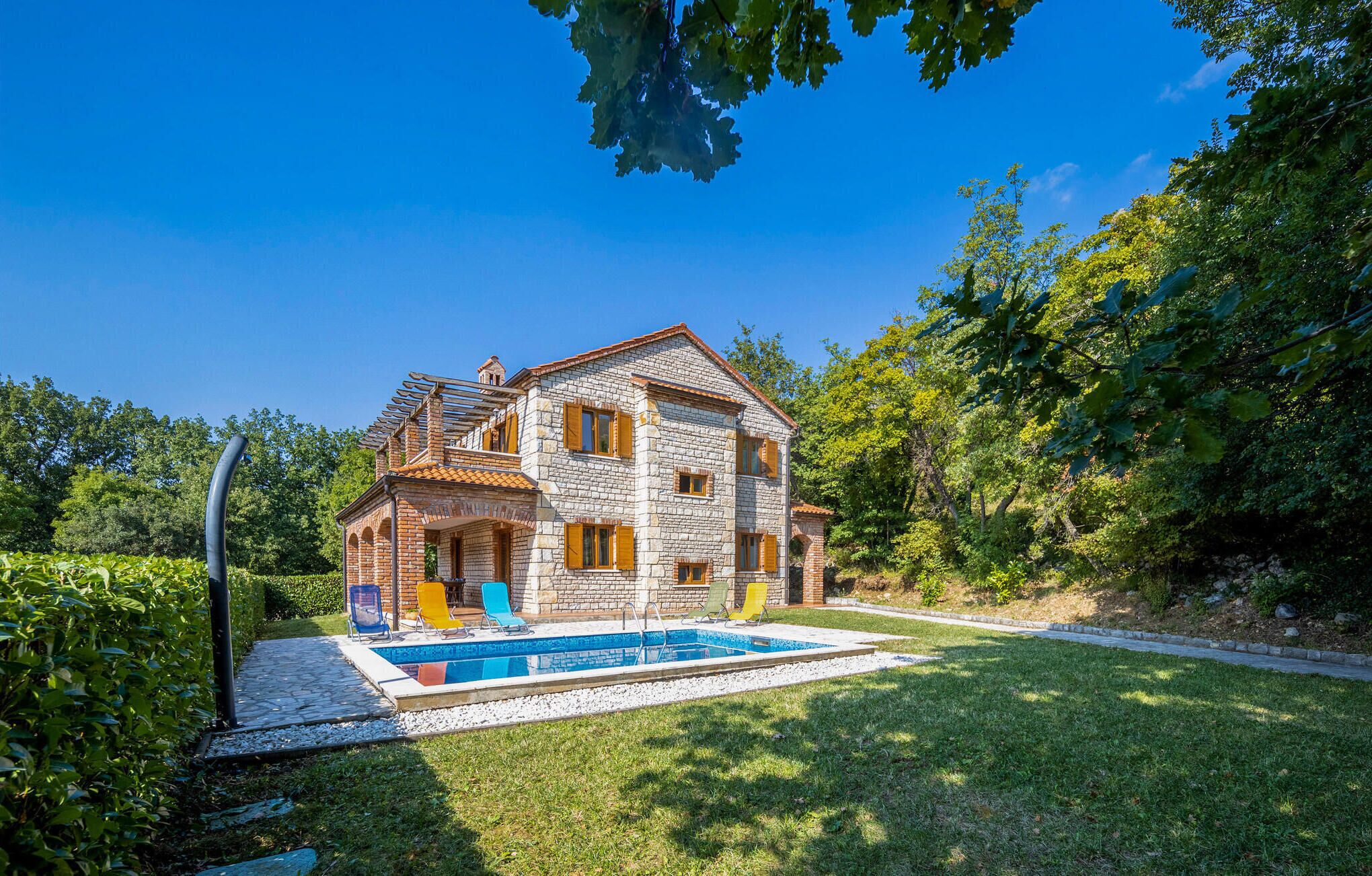 A two-story stone house with a swimming pool, surrounded by lush greenery.