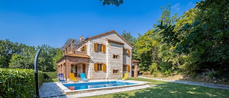 A two-story stone house with a swimming pool, surrounded by lush greenery.