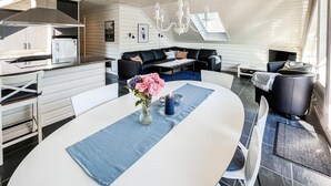 Eine moderne Küche mit weißen Schränken und einem eingebauten Herd. Ein Essbereich mit einem weißen Tisch, blauen Untersetzern und einer Vase mit Blumen. Im Wohnbereich stehen ein schwarzes Sofa und ein schwarzer Sessel.