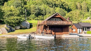 Eine Holzhütte mit einem Balkon, ein Steg mit einem Boot und ein bewaldeter Hügel.