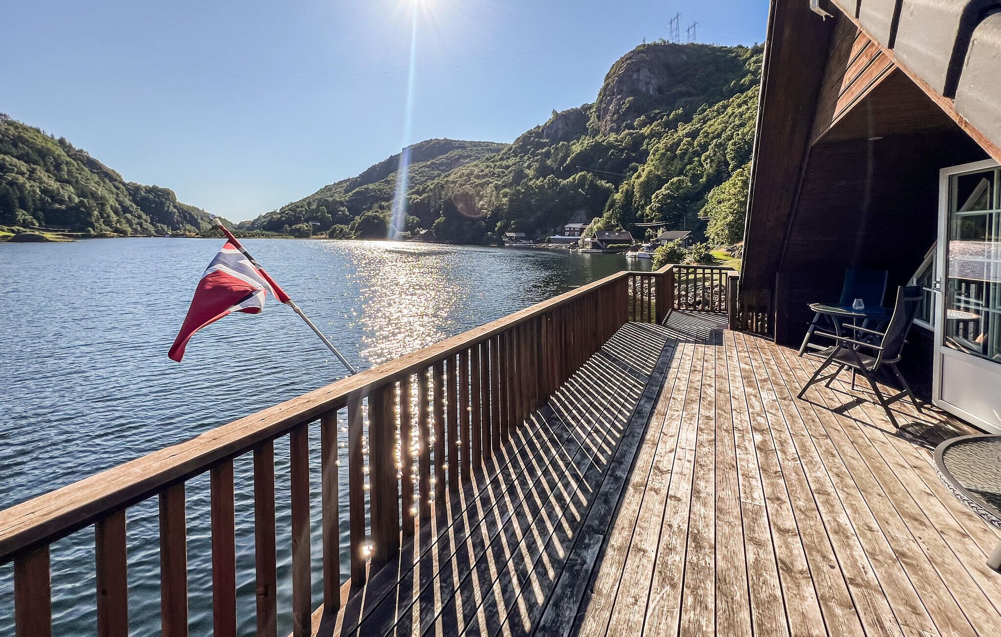 A wooden deck with a railing overlooking a lake, a flag, and a view of a mountainous landscape.