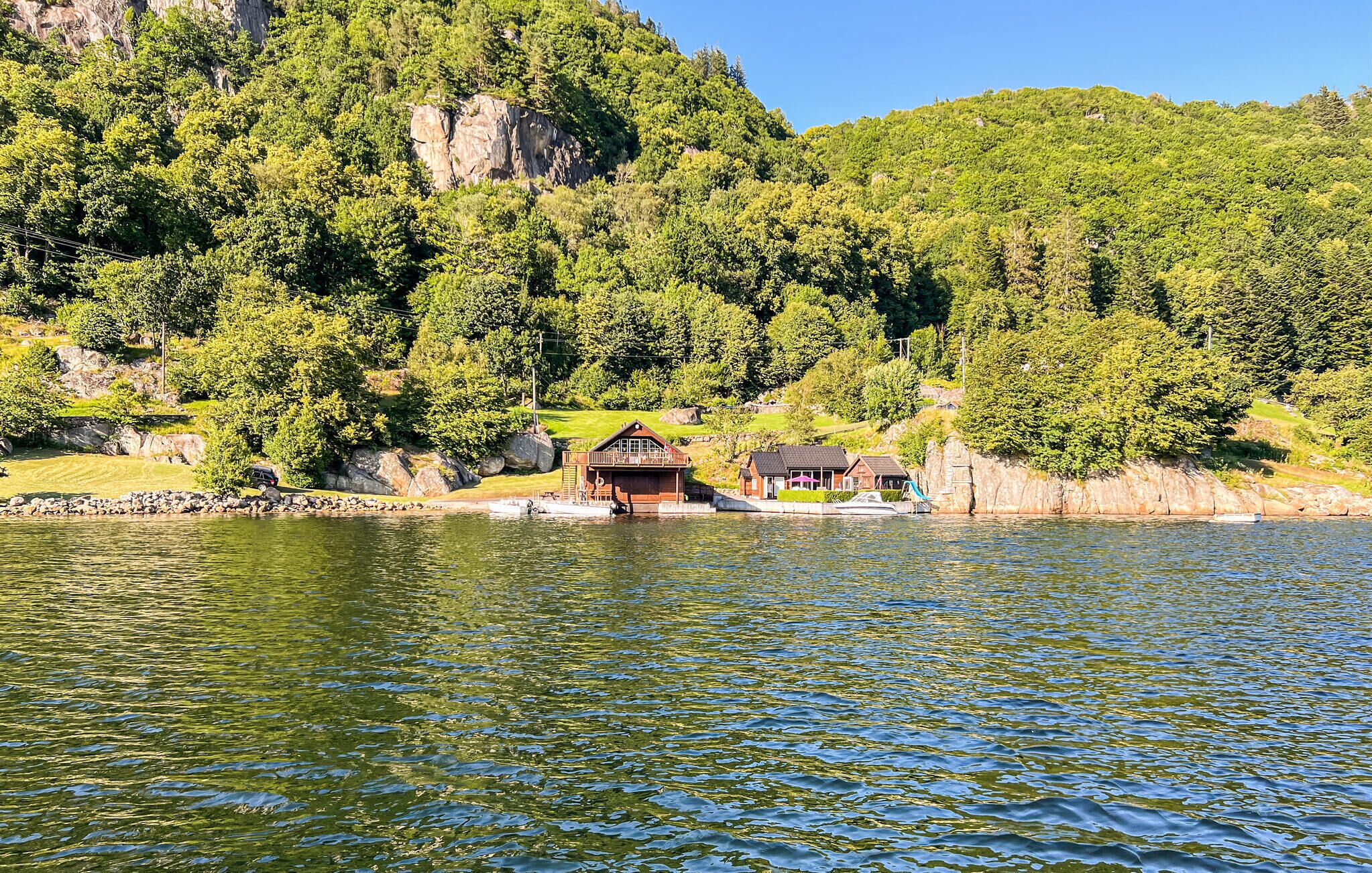 Eine Holzhütte am Seeufer mit Steg, umgeben von dichtem Wald und felsigen Klippen.