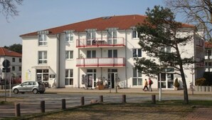 A multi-story building with a white facade and red roof tiles. There are balconies on the second floor, and a car is parked in front.