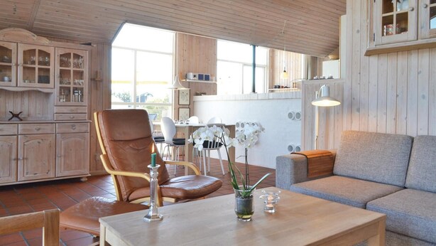 En hyggelig stue med træloft, en læderarmstol i brunt, en grå sofa og et spisebord med et stearinlys og en orkidévase.