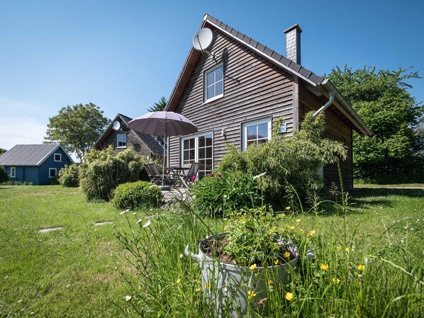 Una casa de madera con antena parabólica, una chimenea y un jardín con flores y plantas.