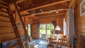 A wooden cabin interior with a loft bedroom, a dining area, and a large sliding glass door leading to an outdoor patio.