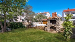 Una cocina exterior de piedra con chimenea, rodeada por un césped cuidado y árboles.
