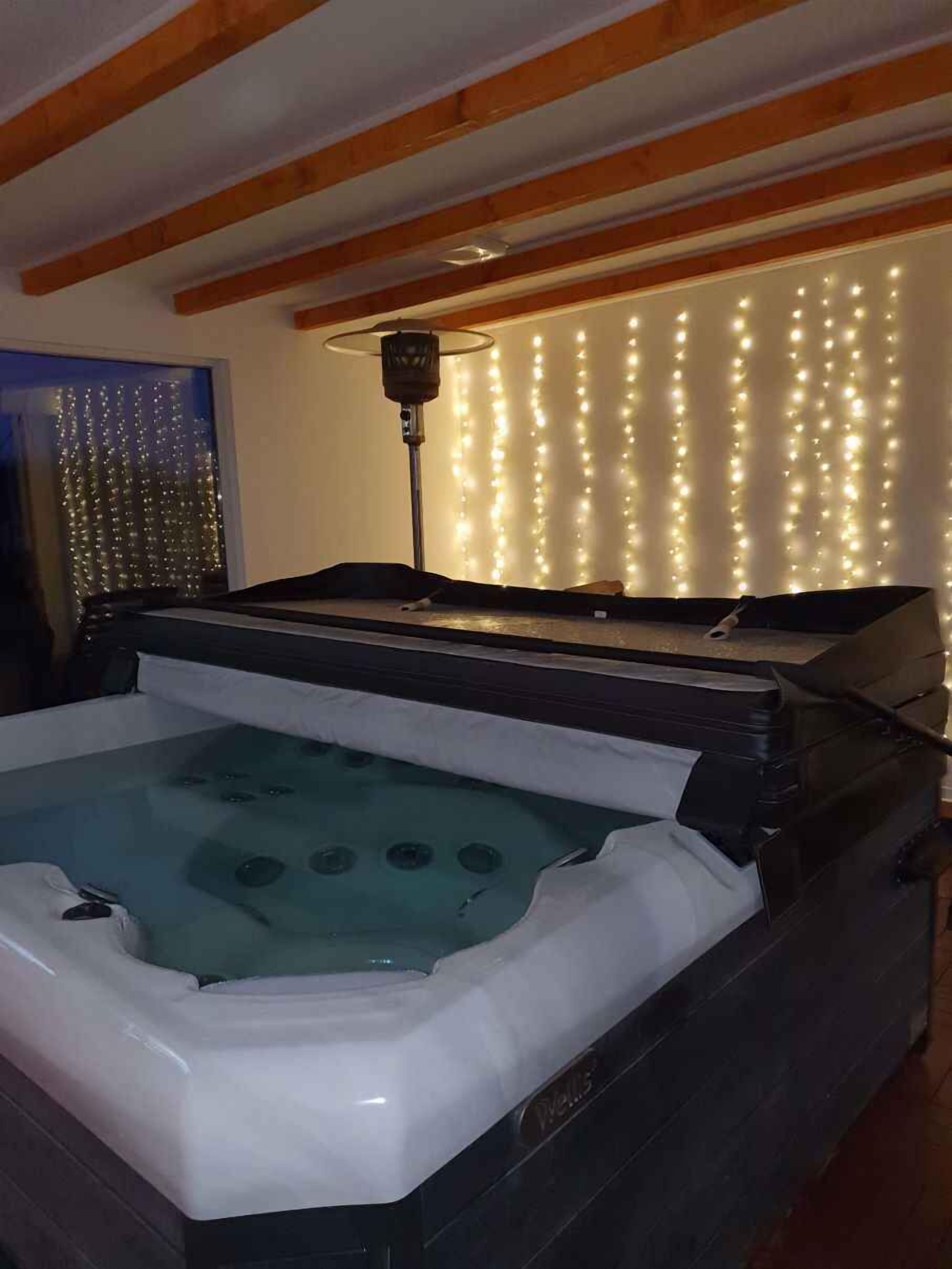 A hot tub in a room with wooden beams, a string of lights on the wall, and a floor lamp.
