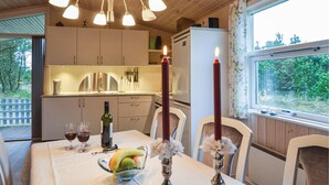 Un área de comedor con una mesa lista para dos personas, una botella de vino y un frutero. Hay velas sobre la mesa, y se ve una ventana con vistas a árboles.