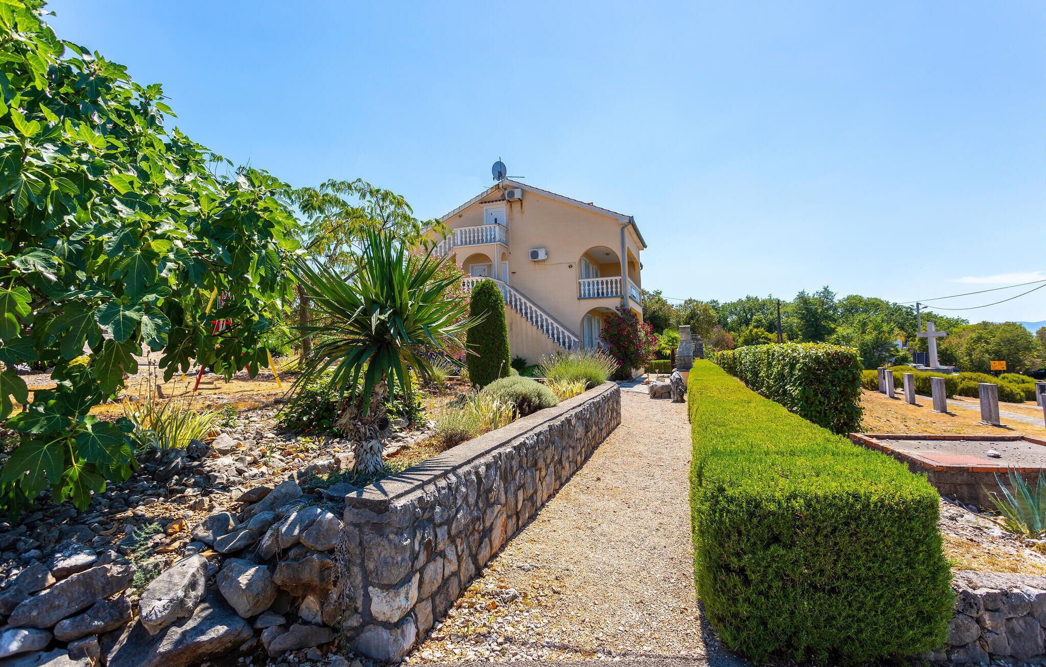 Eine Villa im mediterranen Stil mit einem gepflasterten Weg, der zu einem verschlossenen Eingang führt, umgeben von üppiger Vegetation und einem gepflegten Garten.