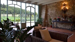 A spacious room with a large window offering a view of a garden, wicker furniture, a stone wall, and a chandelier. - Holiday apartment with pool (Castelluccio Capolona)