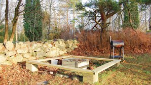 A rustic outdoor grill setup with a stone wall and wooden bench in a wooded area.
