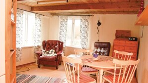 A cozy room with a wooden ceiling, a dining table with chairs, a red leather armchair, a wooden cabinet, and patterned curtains.