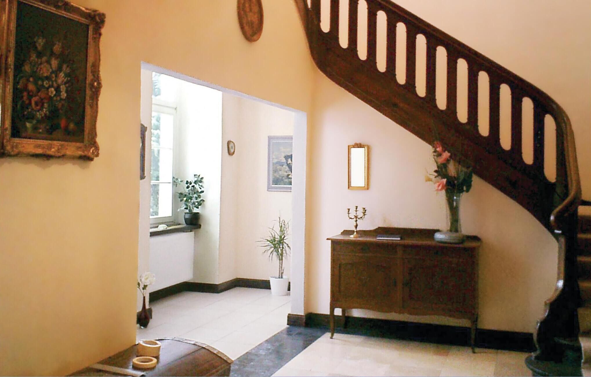 Un couloir avec un escalier en bois, une plante en pot, un miroir et un meuble.