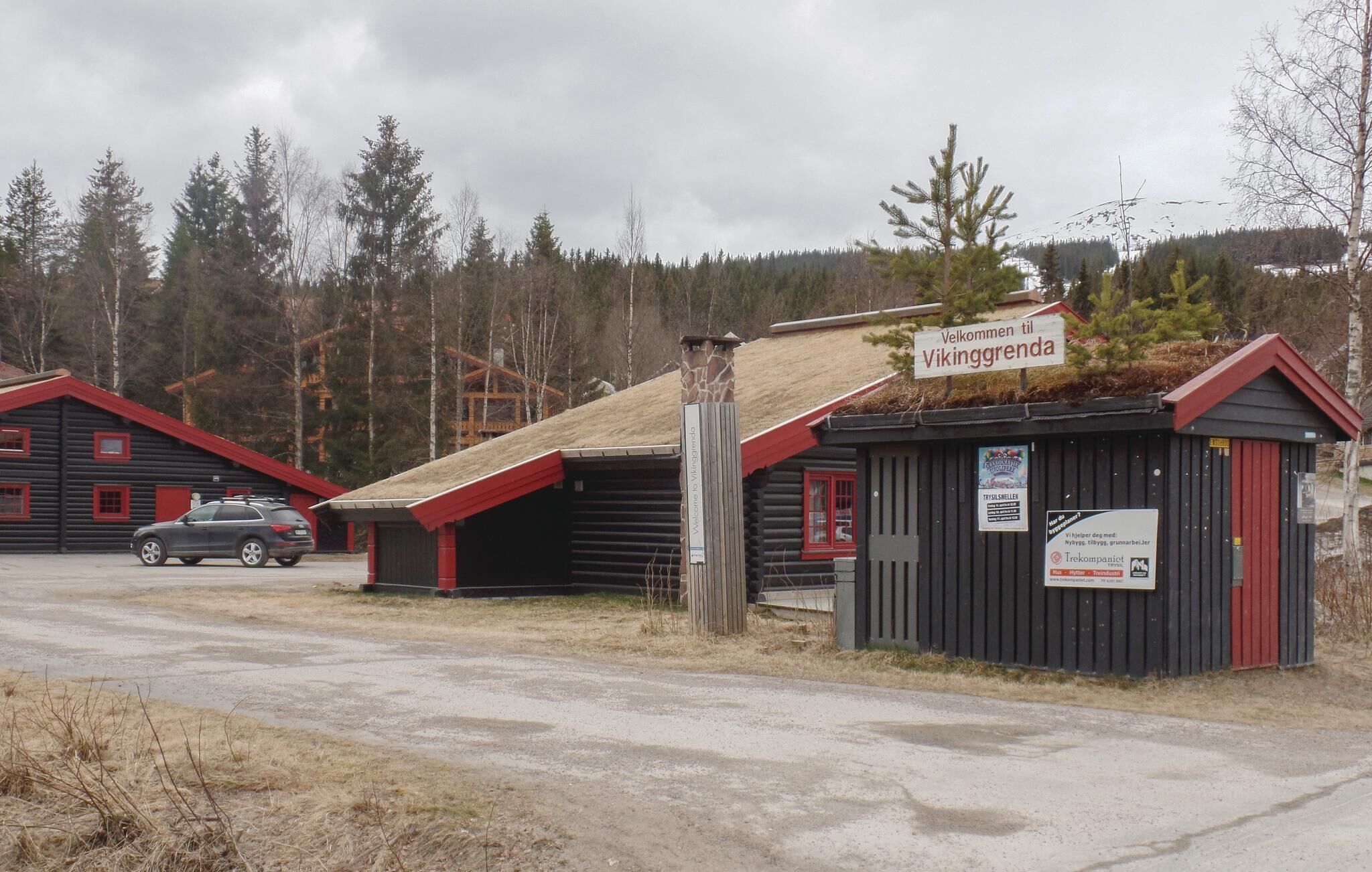 A rustic cabin with a 'Welcome to Vikinggrenda' sign, a small wooden structure, and a car parked nearby.