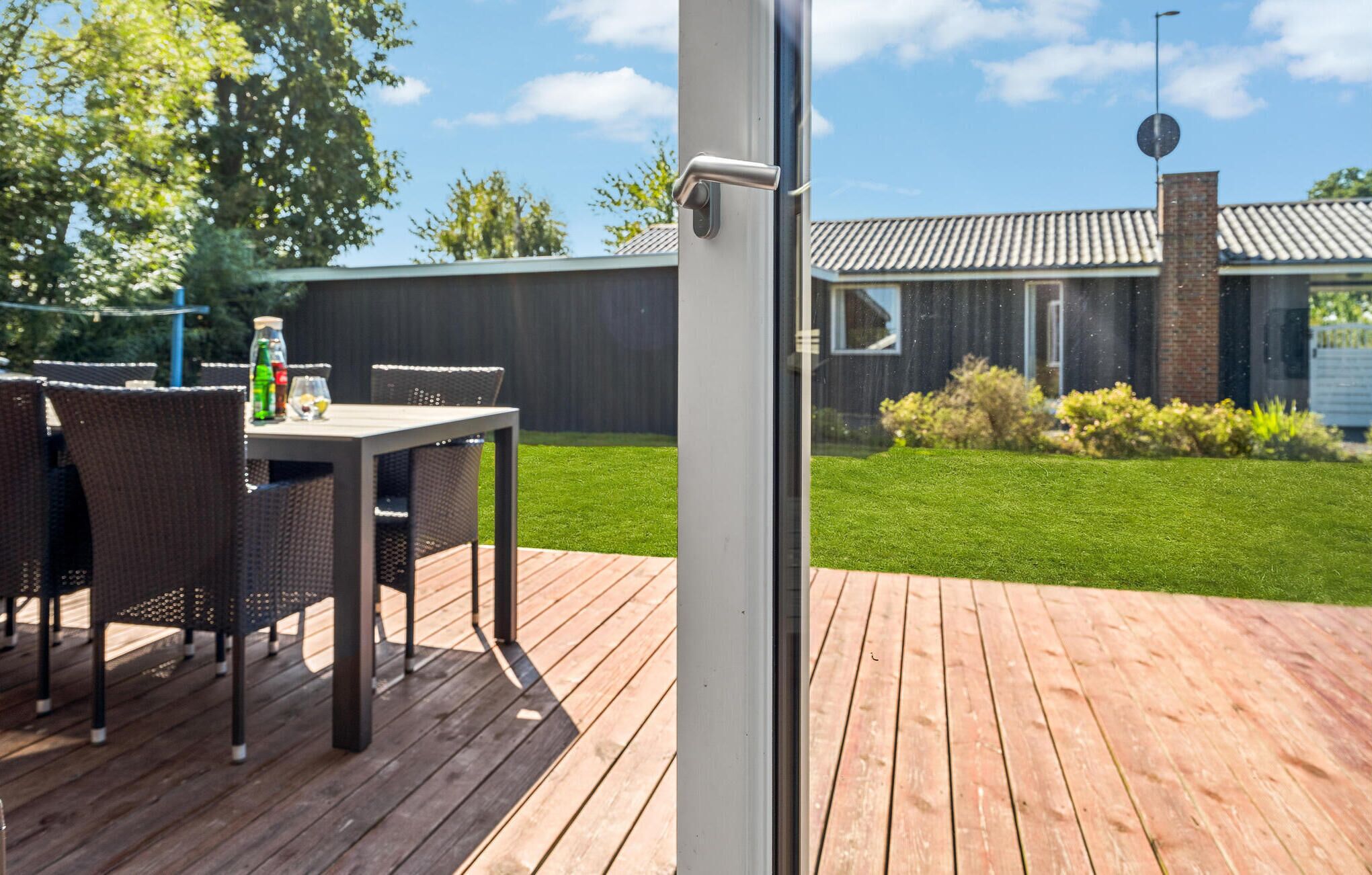 Een terras buiten met een tafel en stoelen, een houten terras en uitzicht op een huis met een bakstenen schoorsteen.