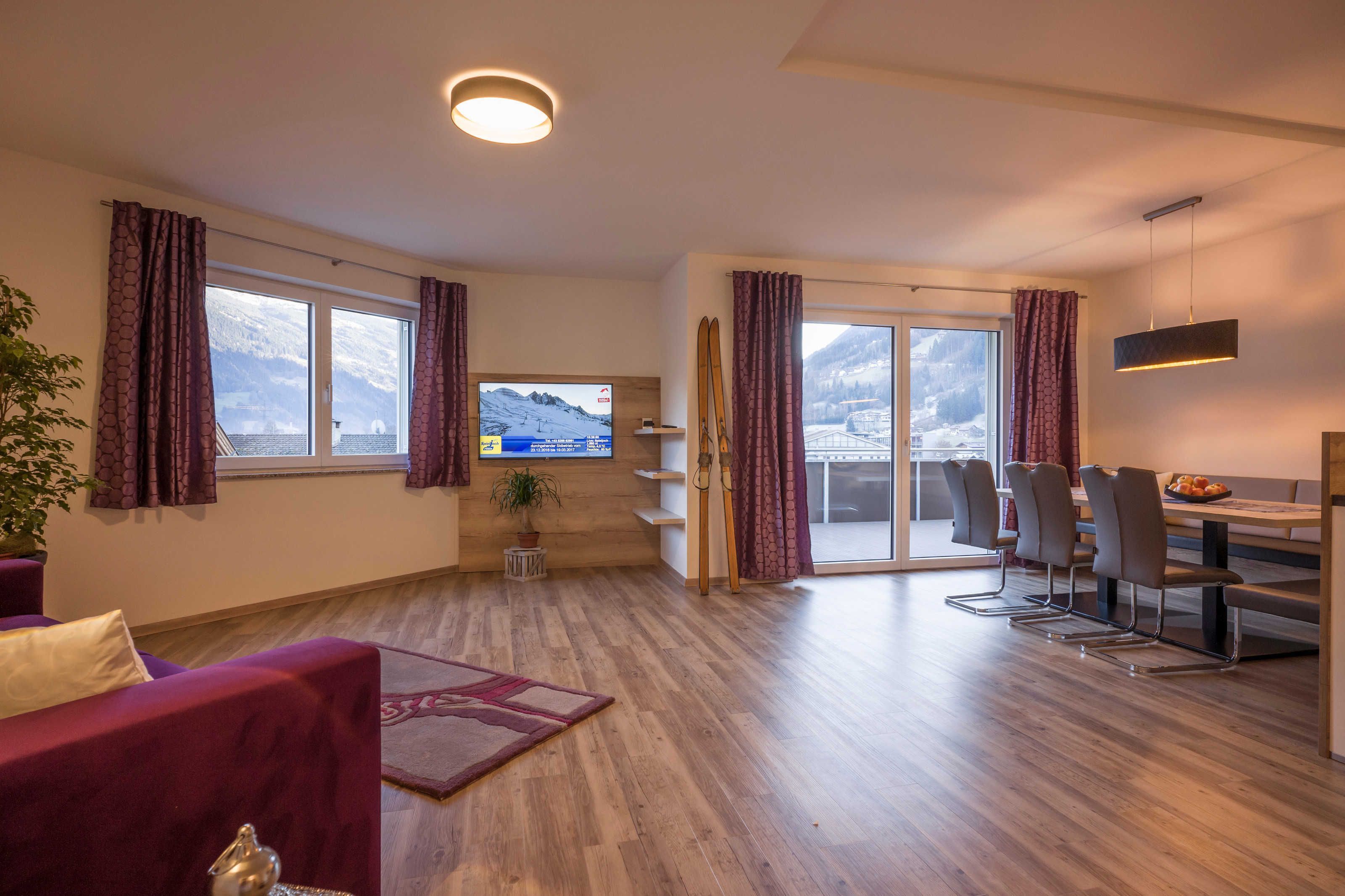 Ein modernes Wohnzimmer mit Fernseher, Holzboden, einer Couch, einem Essbereich mit Stühlen und Blick auf einen verschneiten Berg durch große Fenster.