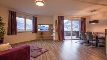 Ein modernes Wohnzimmer mit Fernseher, Holzboden, einer Couch, einem Essbereich mit Stühlen und Blick auf einen verschneiten Berg durch große Fenster.