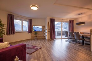 Una sala de estar moderna con una televisión, piso de madera, un sofá, un comedor con sillas y vistas a una montaña nevada a través de grandes ventanales.