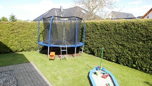 A trampoline with a safety net, a small play area with toys, and a hedge in a backyard. - Vacation home Sellin, child-friendly, barbecue, terrace, WLAN - Holiday home (Karlshagen)