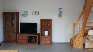 A living room with a TV, wooden cabinets, a staircase, and artwork on the walls. - Vacation home Sellin, child-friendly, barbecue, terrace, WLAN - Holiday home (Karlshagen)