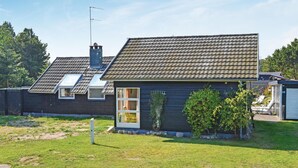 Une maison de plain-pied avec un toit en pente, une cheminée bien visible et un petit jardin devant, composé d’une pelouse et de quelques arbres.