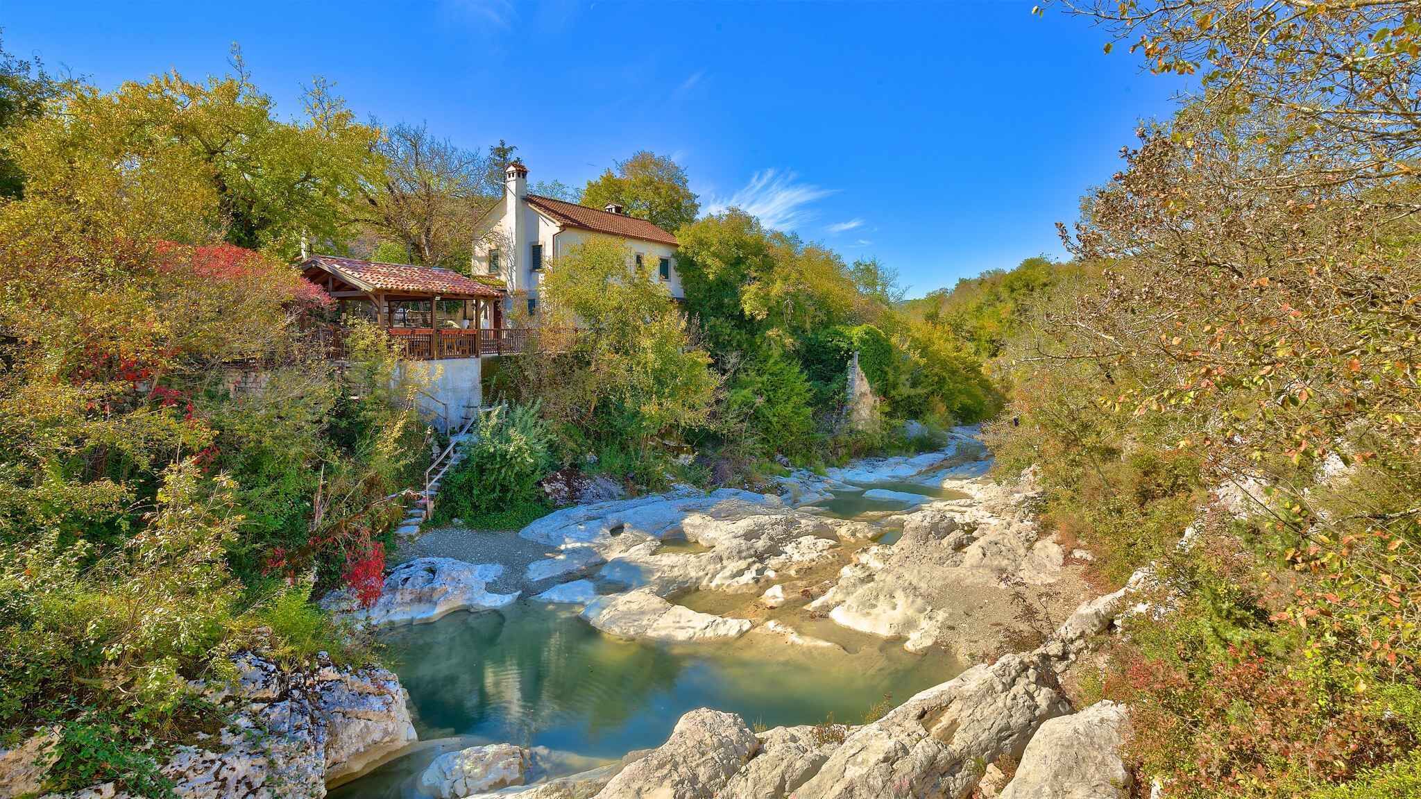 A rustic house with a stone staircase leading to a wooden deck, surrounded by lush greenery and a clear stream.