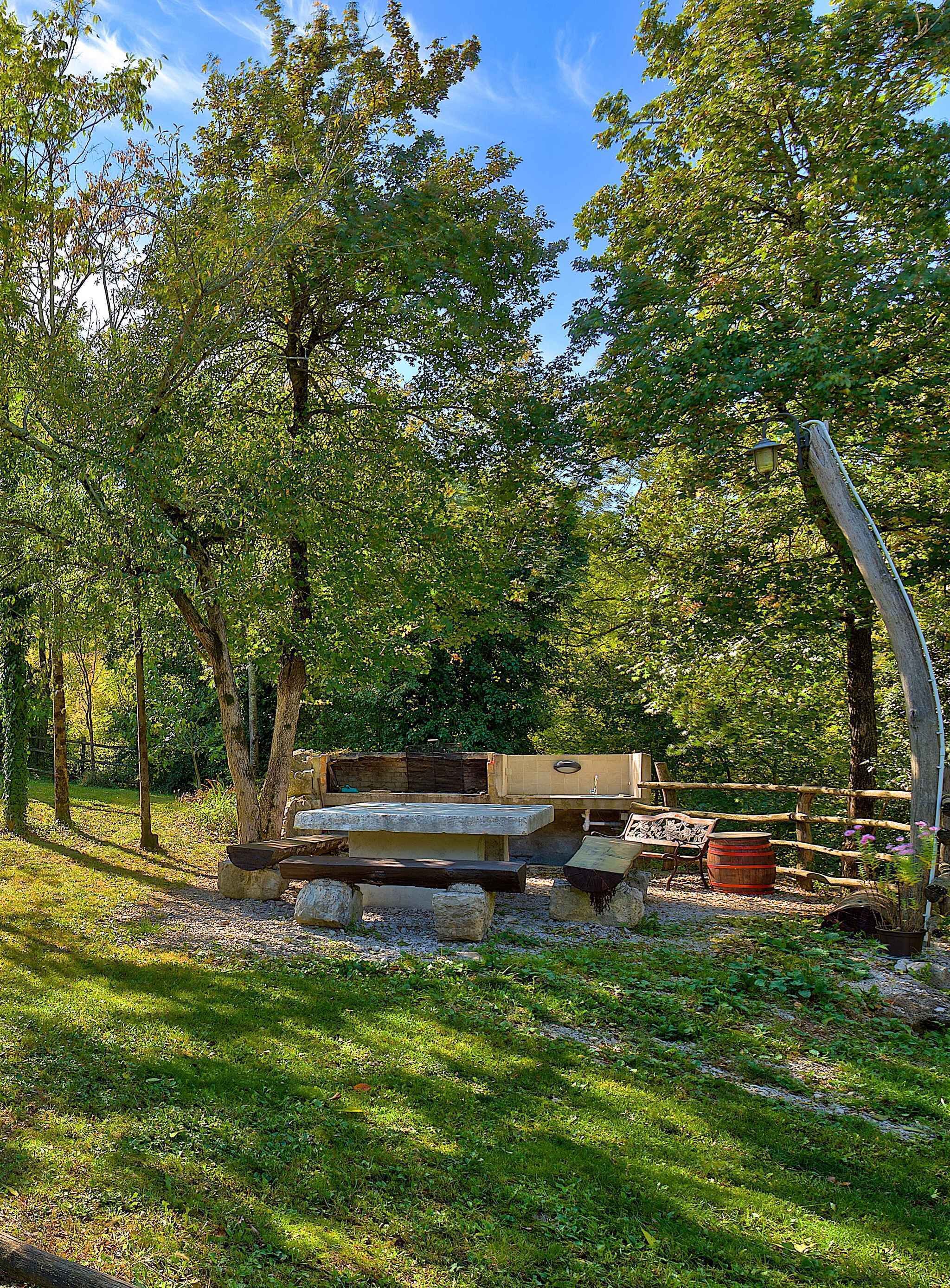 A rustic outdoor seating area with stone benches and a wooden fence, surrounded by lush greenery and trees.