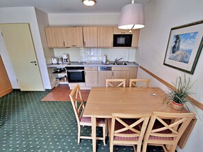 A compact kitchen with wooden cabinets, a microwave, a stove, and a sink. There is a dining table with chairs and a potted plant.