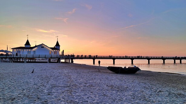 Ein Strand mit einer Mole, einem Boot und einem weißen Gebäude mit zwei Türmen.