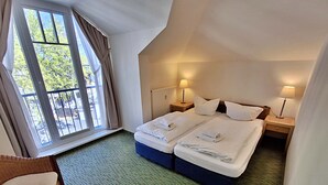 Ein Hotelzimmer mit zwei Betten, einem Fenster mit Vorhängen, einem Sessel und zwei Nachttischlampen.
