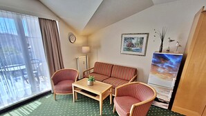 Ein Hotelzimmer mit grünem, gemustertem Teppich, einer Couch, zwei Sesseln, einem kleinen Tisch mit einer Pflanze, einer Uhr, einem gerahmten Bild und einem Fernseher.