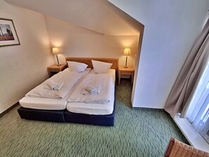 A hotel room with two beds, each with a pillow and a folded towel. There are two bedside tables with lamps, a framed picture on the wall, and a window with curtains.
