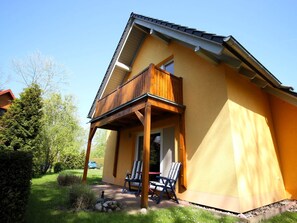 Une maison à deux étages avec un balcon en bois, un petit patio avec deux chaises et un ciel d’un bleu limpide.