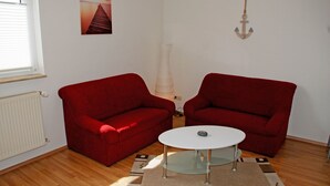 Une pièce avec deux canapés rouges, une table basse blanche, un tapis à motifs, un radiateur blanc, un tableau accroché au mur et une ancre fixée au mur.