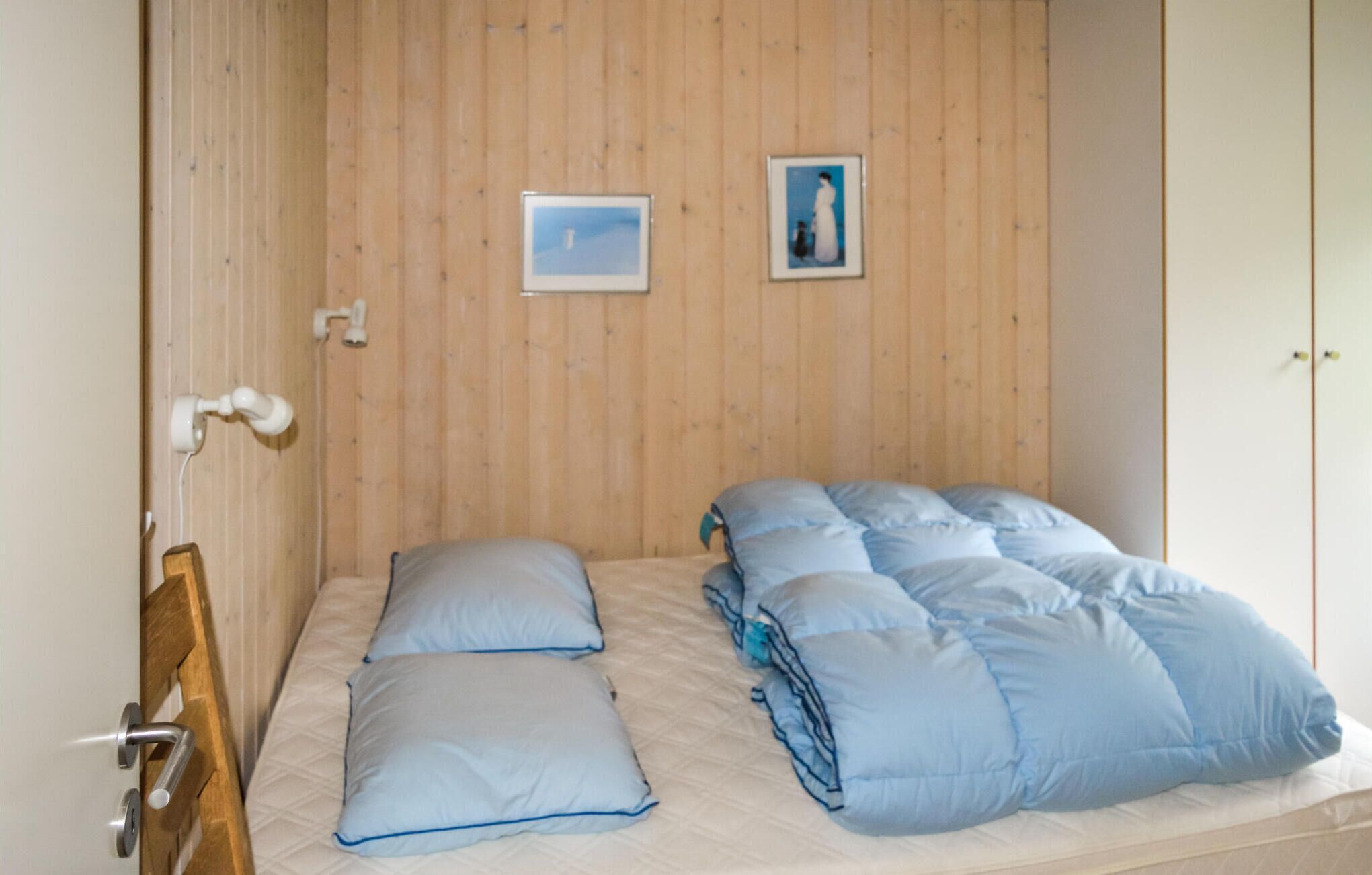 Een kamer met een enkel bed, een houten wand, een deur met een hendel en twee ingelijste foto's.