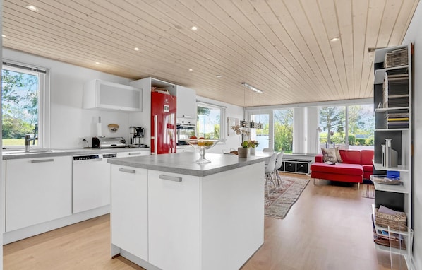 Une cuisine moderne avec des meubles blancs, un réfrigérateur rouge et un îlot central sur lequel est posé un panier de fruits.
