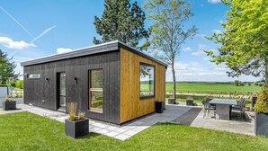 Un edificio moderno de dos pisos con fachada oscura y detalles de madera, que cuenta con amplios ventanales y un patio con mobiliario exterior.
